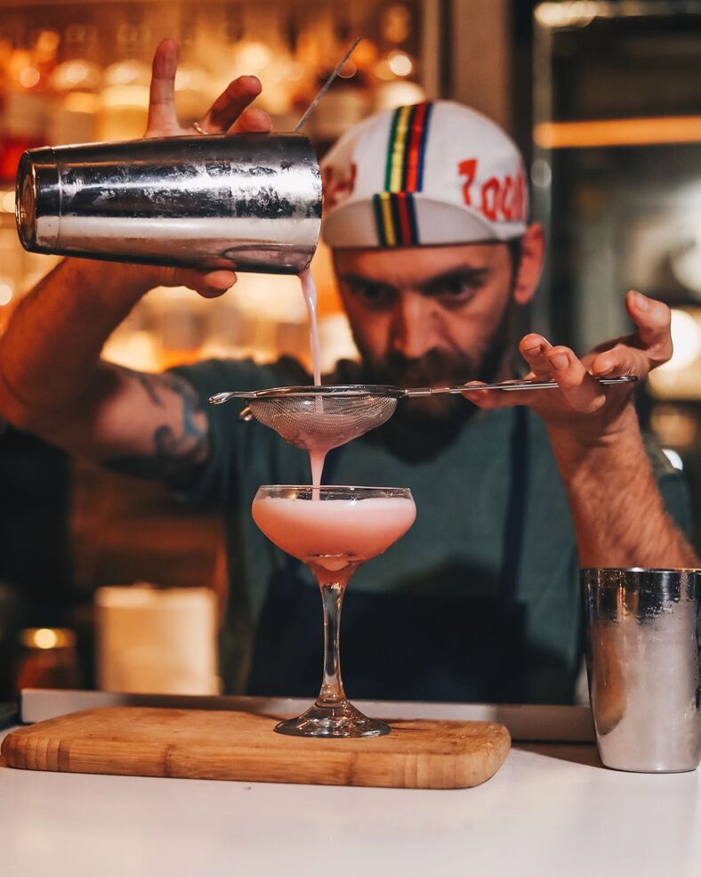 Bearded bartender strains a pink beverage into a cocktail glass. 