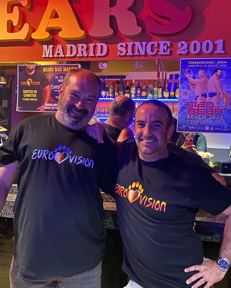 Two scruffy middle-aged men in Eurovision t-shirts pose with arms around each other in front of the Bears Bar sign. 
