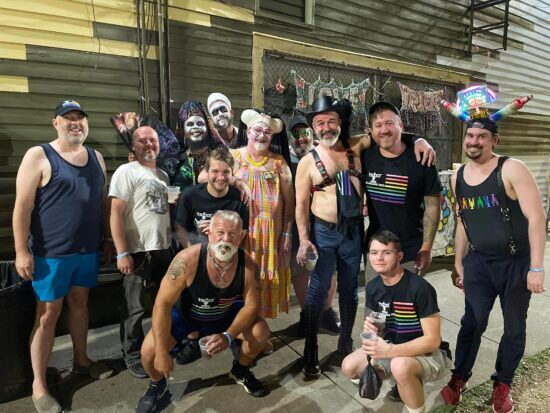 A gaggle of shirtless daddies in leather and rainbow pride shirts, festively dresses bearded guys, and two Sisters of perpetual Indulgence pose in front of the Phoenix 