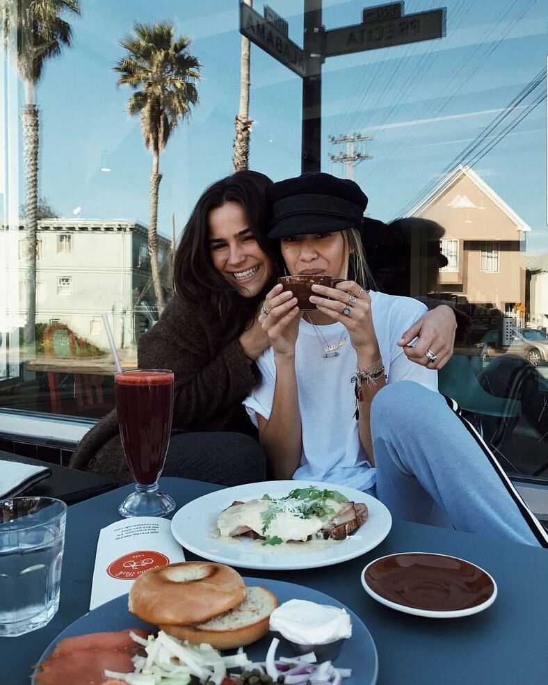 Two women pose for a photo at their tables filled with brunch items and coffee on a sunny day Precita Park Cafe.