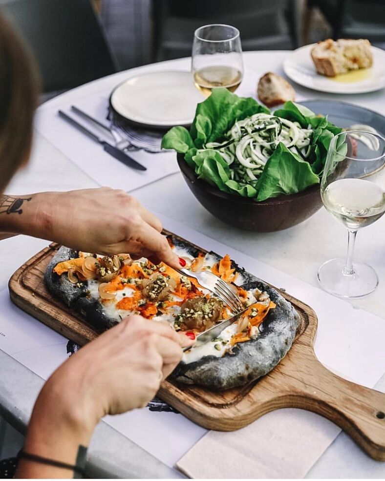 A person cuts into a black squid ink pizza covered in colorful orange toppings. The meal is accompanied by a fresh green salad and a crisp white wine. 