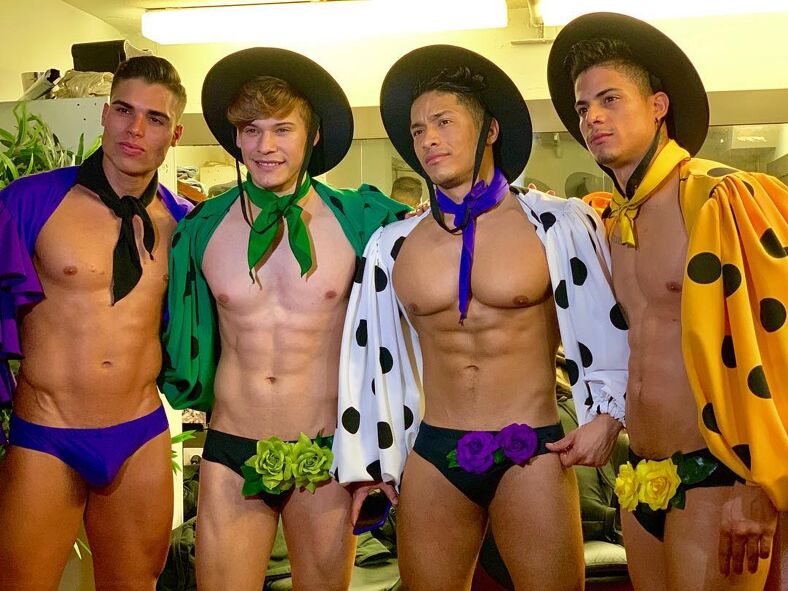 Four shirtless hunky Spaniards look into the camera adorned with skimpy briefs, bulging packages, multi-colored polka-dot capes tied around their necks, and black brimmed Sierras hats.