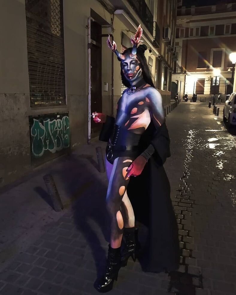 Alien-looking club kid with horns, muliti-colored and full-body makeup poses with a cigarette in the streets of Malasaña