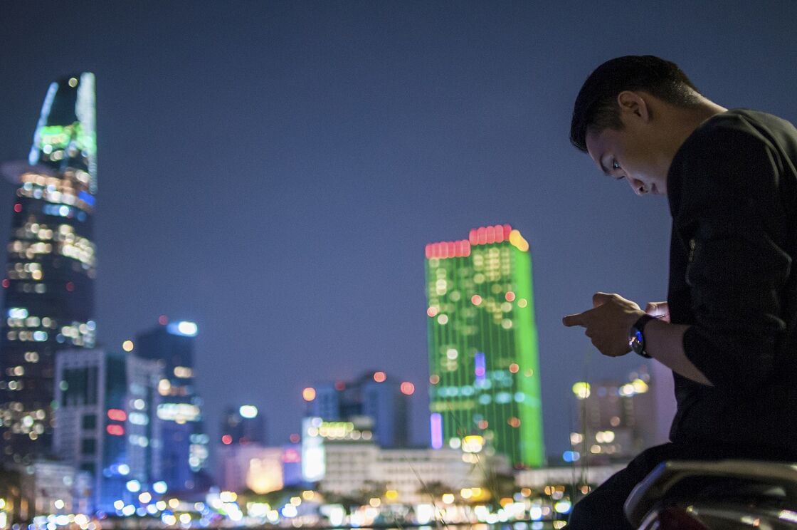 Man texts on his phone with the bright lights of Saigon in the background.