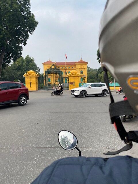 Point of view of a passenger riding on the back of a motorbike with oncoming traffic and the canary yellow Vietnamese presidential palace in sight.