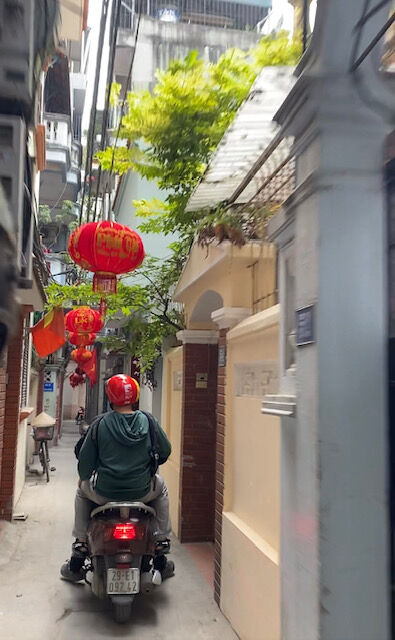 Motorbike and passengers zoom through a narrow alley with hanging red lanterns, foliage, and the red Vietnamese flag. 