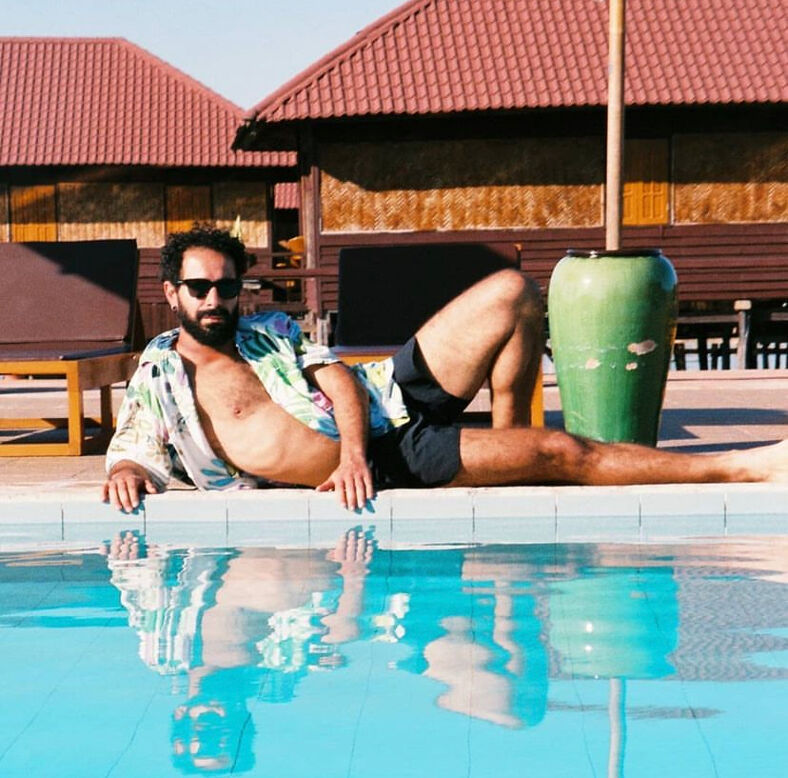 Ouissam poses with sunglasses and an open shirt by the pool with a backdrop of terracotta shingled buildings. 