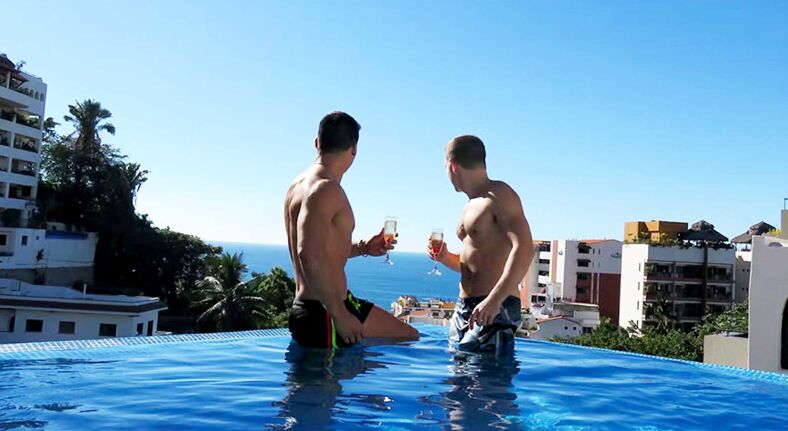 Two shirtless men holding champagne flutes in a pool.
