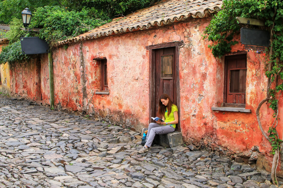 A woman reads a book in the doorway of a humble historic red building with a tiled roof on a cobble stone street in Uruguay.