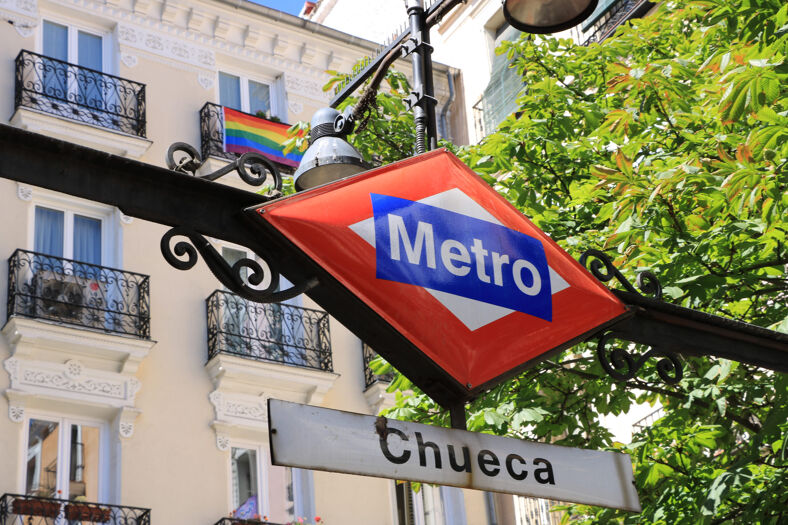 Chueca metro station sign with a rainbow flag hanging off a balcony in the background.