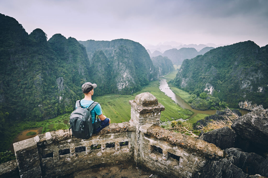 Man with a backpack sits on an ornate stone ledge overlooking a green river valley in Vietnam.