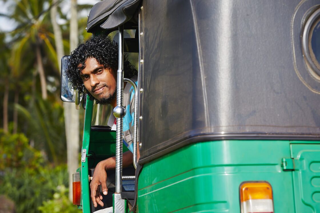 A handsome Sri Lankan man looks back at the viewer from the driver's side of a Tuk Tuk.