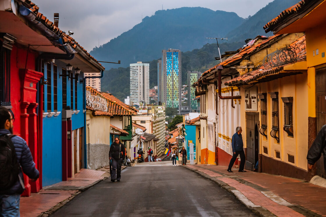 Looking down a street in Bogota with brightly colored buildings, people walking, and in the distance a multi-colored skyscraper and green mountains.