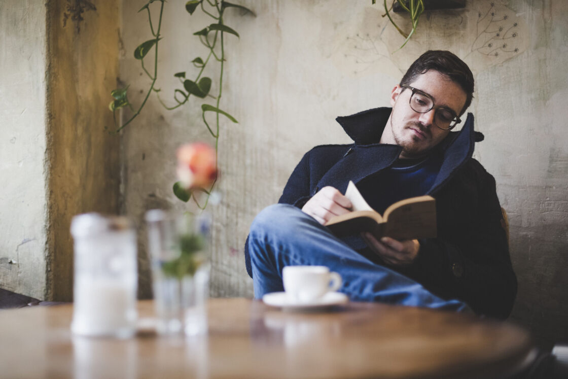 Man in peacoat reads a book in a quaint coffee shop with hanging vines in the distance