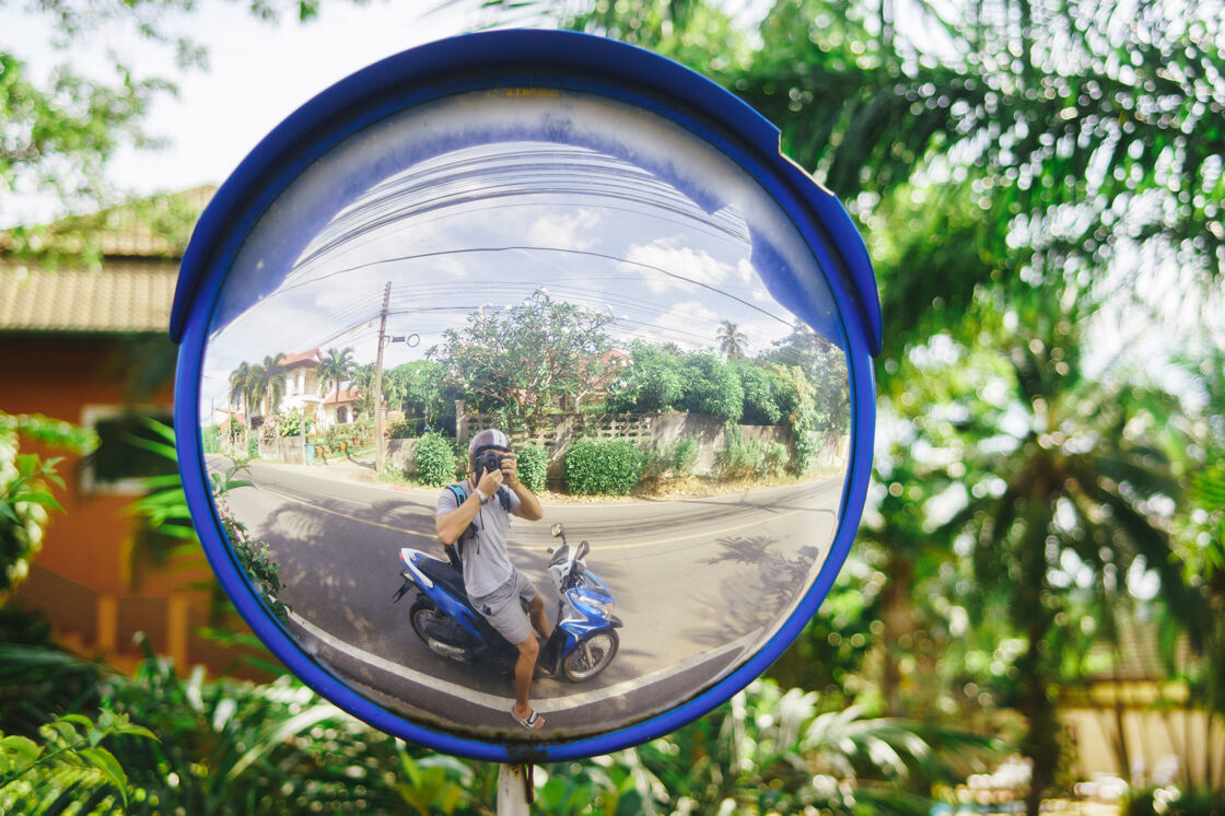 Man on blue scooter takes a photo of himself in a convex traffic mirror revealing a landscape of lush plant life around a house and concrete fence in Taiwan.