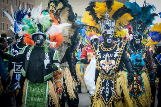 Black members of the Krewe of Zulu  dressed in colorful headdresses and grass skirts march through the streets of New Orleans.