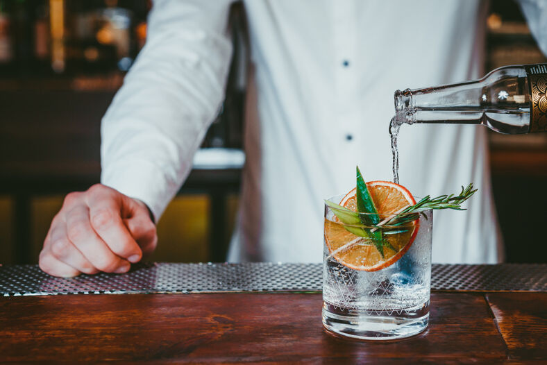 Bartender pours tonic water into a glass of gin decorated with a candied orange slice and a sprig of rosemary.