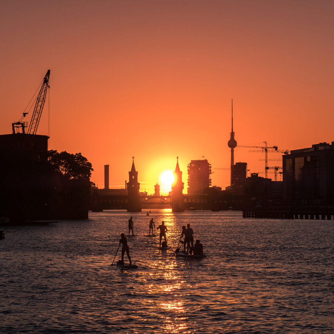 Paddle-boarders paddle towards sunset on the river Spree in Berlin with a bridge resembling a castle, towering cranes, and the famous Berlin TV tower in the background.