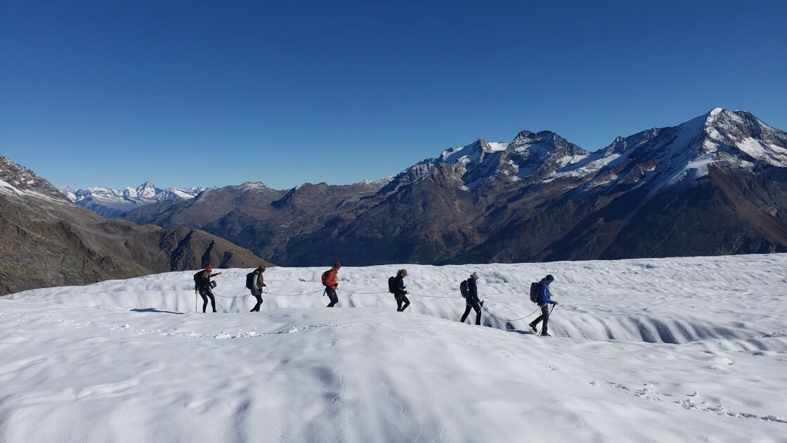 Six people from a distance traverse a glacier with snowcapped mountains in the background. 