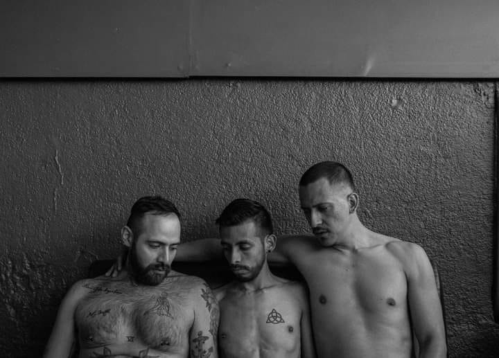 Three shirtless handsome tattooed men peer at each other's crotches which are outside of the frame.  