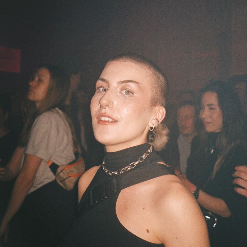 A person with a shaved head, facial piercings, and hazel eyes peers at the camera at Under Bron in Stockholm.