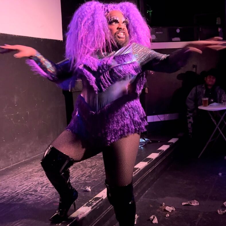Coco Buttah lip-sings at Dollz in a purple wig and fringed body suit. 