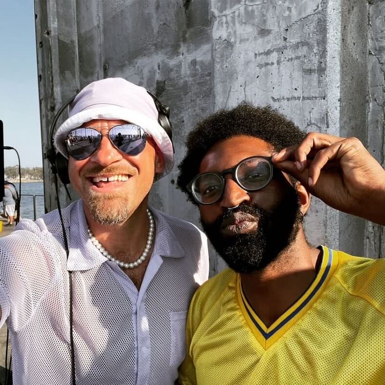 DJ Nate Manic in a white mesh shirt and sunglasses and Dj Charles Hawthorne in canary yellow embrace and smile at the camera.