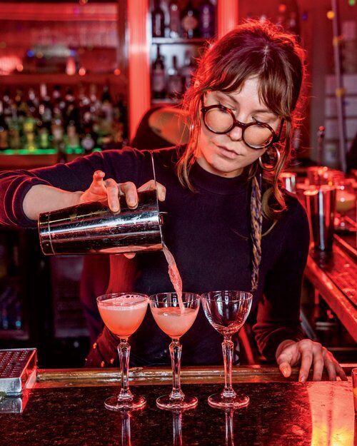 A bartender in braids and round frame glasses pours cocktails.