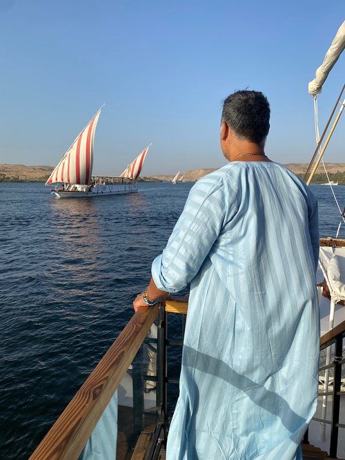 A man in a light blue caftan looks onto the water and passing boats on a Nile river cruise in Egypt.