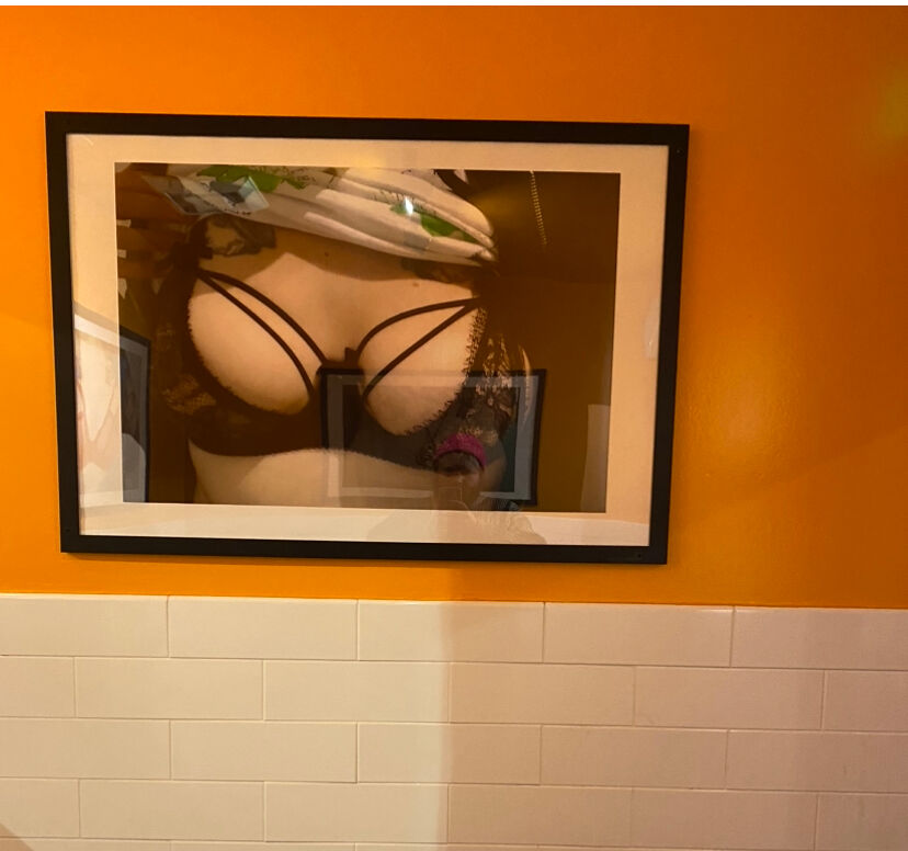 A framed picture of breasts in a sexy lingerie bra against an orange wall in Mother's gender-neutral bathroom.