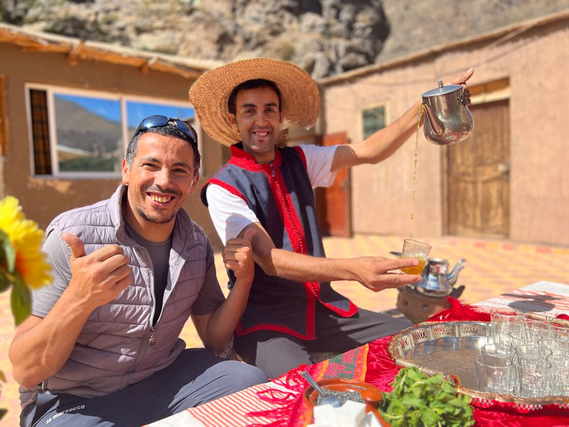 A man with thumbs-up sits next to another who is pouring tea in a arid Moroccan village.