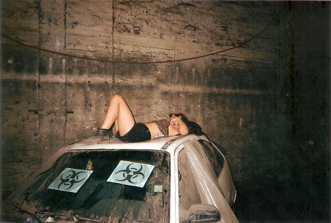 A raver in sunglasses lays on top of a dusty white car with biohazard signs in the window. 