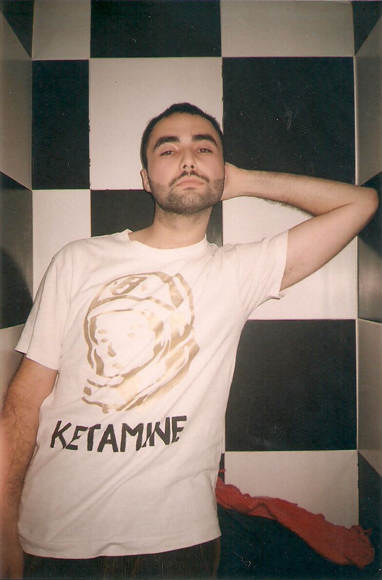 A good-looking guy with subtle white eye makeup poses in front of a room of black and white checkered tiles. His white shirt says "ketamine" under an astronaut.