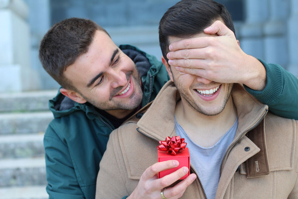 Man surprising boyfriend with red gift box in hand.