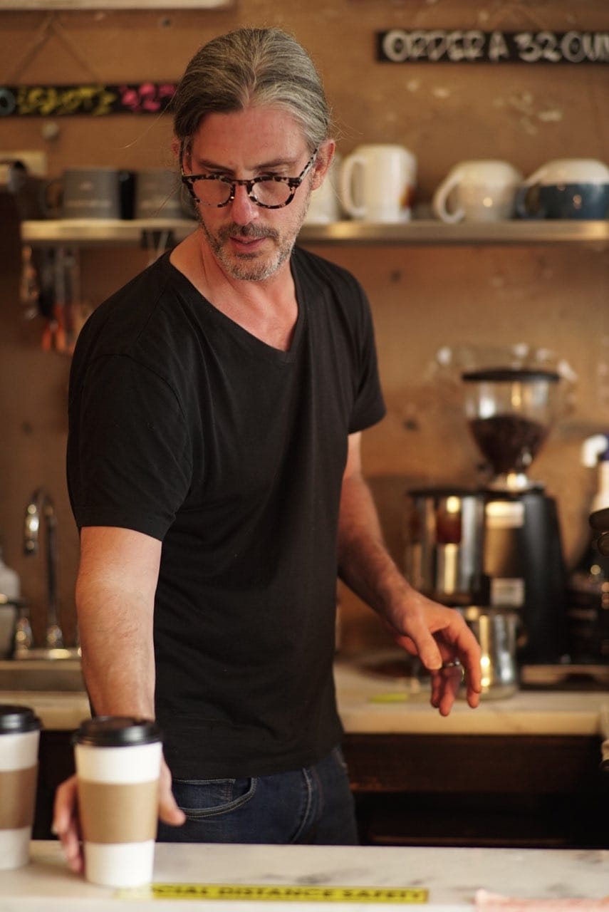 A handsome middle-aged gentleman in tortoise shell glasses with salt and pepper hair puts a coffee on the bar. 