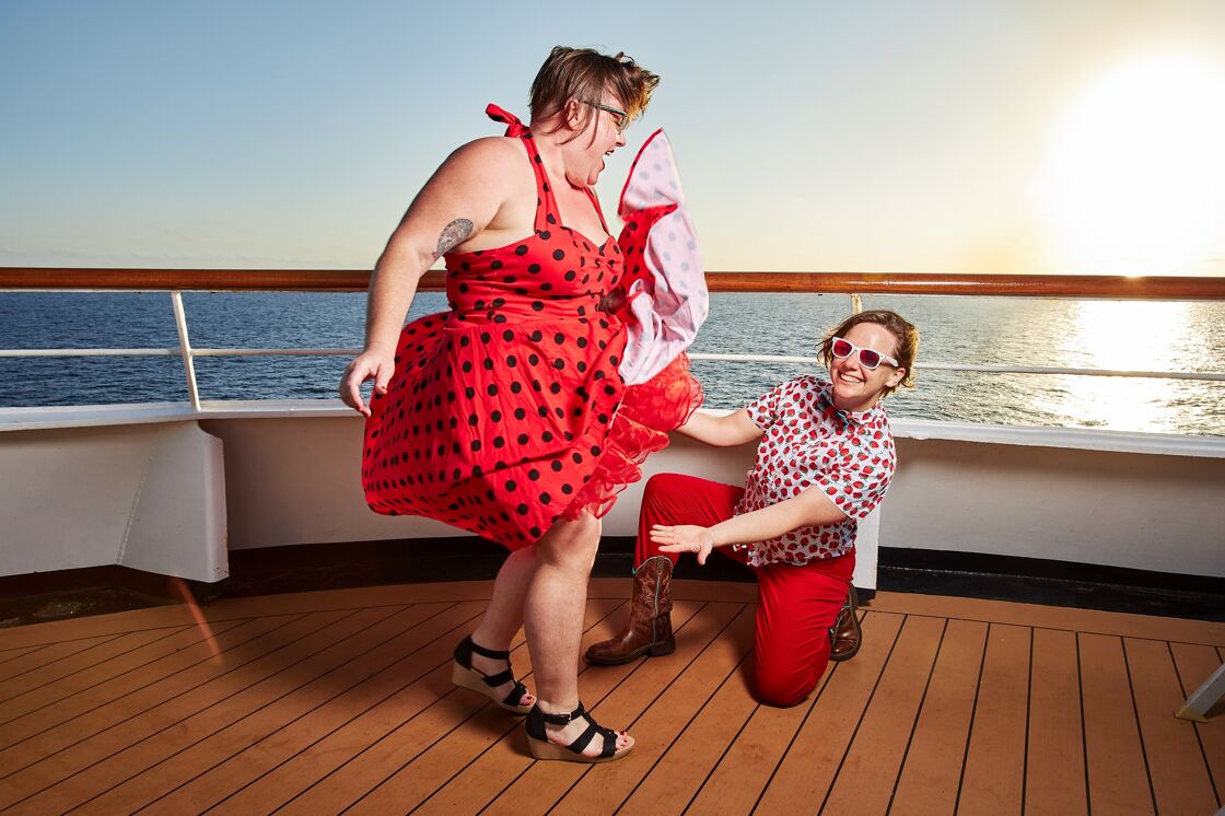 A woman in a red polka dot dresses dances twirling her skirt while her dancing partner in brown cowboy boots, red pants, and a red strawberry shirt smiles on one knee. 
