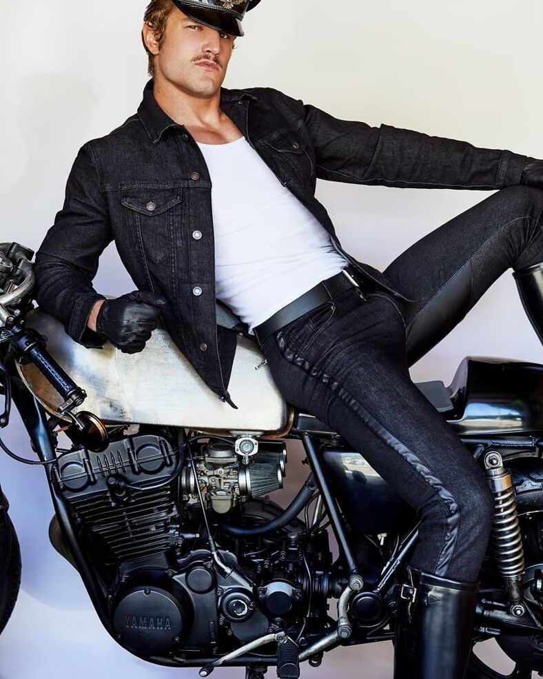 A handsome man dressed in jeans and leather chaps leans against a motorcycle.