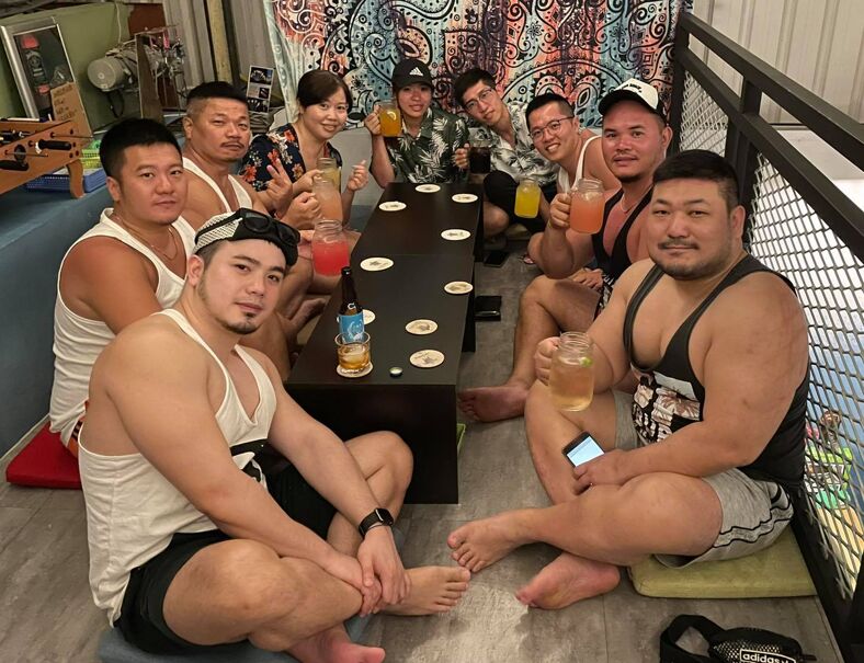 A group of muscle bears and their admirers hold up their glasses around a table at Bear Junkies.