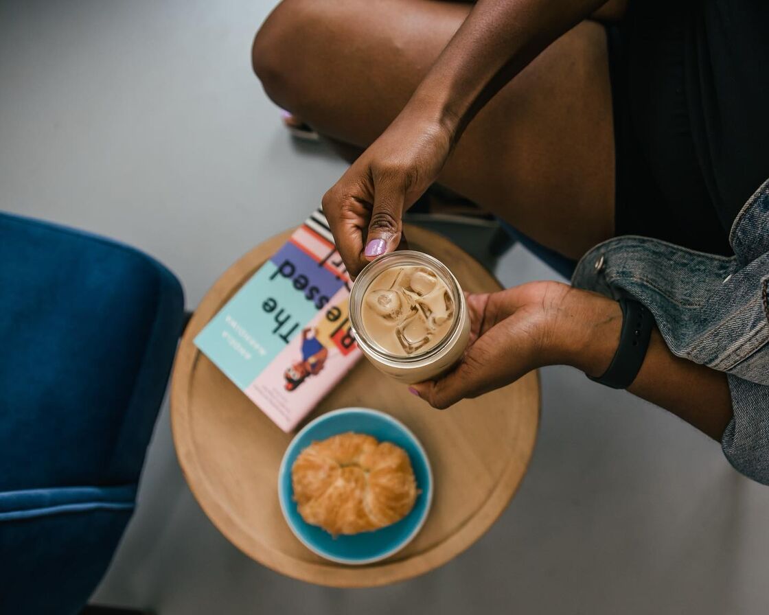 A woman holds an iced coffee drink above a colorful book and croissant. 