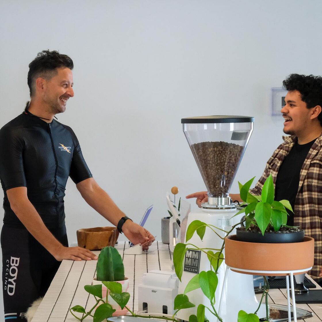 A coffee customer in  black lycra cycling gear pays for his coffee as the hip  barista in plaid smiles.