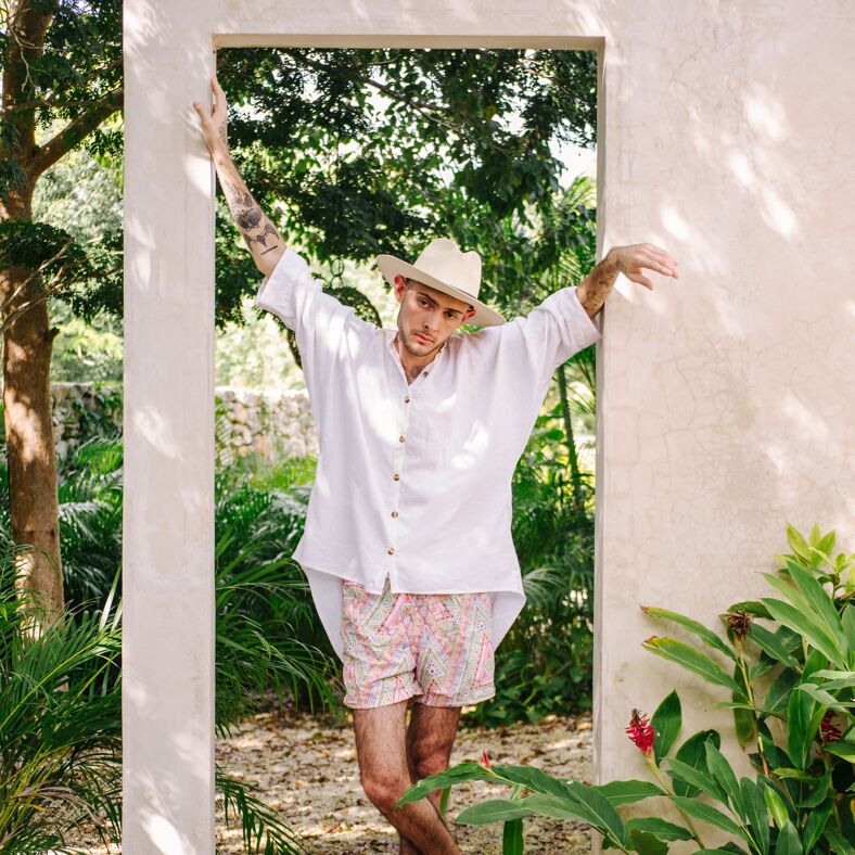 A stylish man leans on an alabaster door frame in a lush garden while wearing an oversized white linen shirt, beige hat, and colorful printed shorts. 