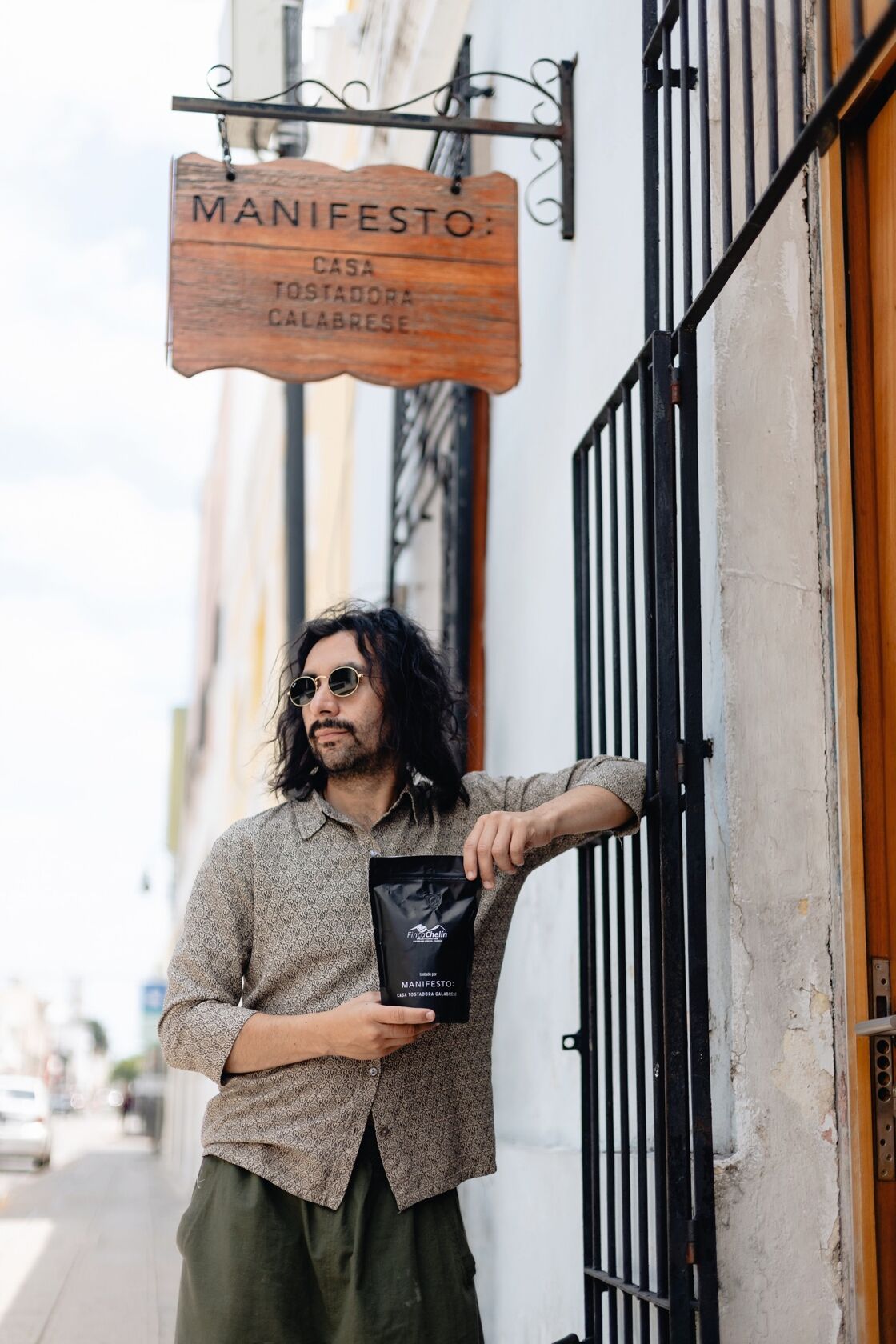 A shaggy man with sunglasses looks away from the camera as he poses with a bag of coffee under the Manifesto Coffee Roasters sign.