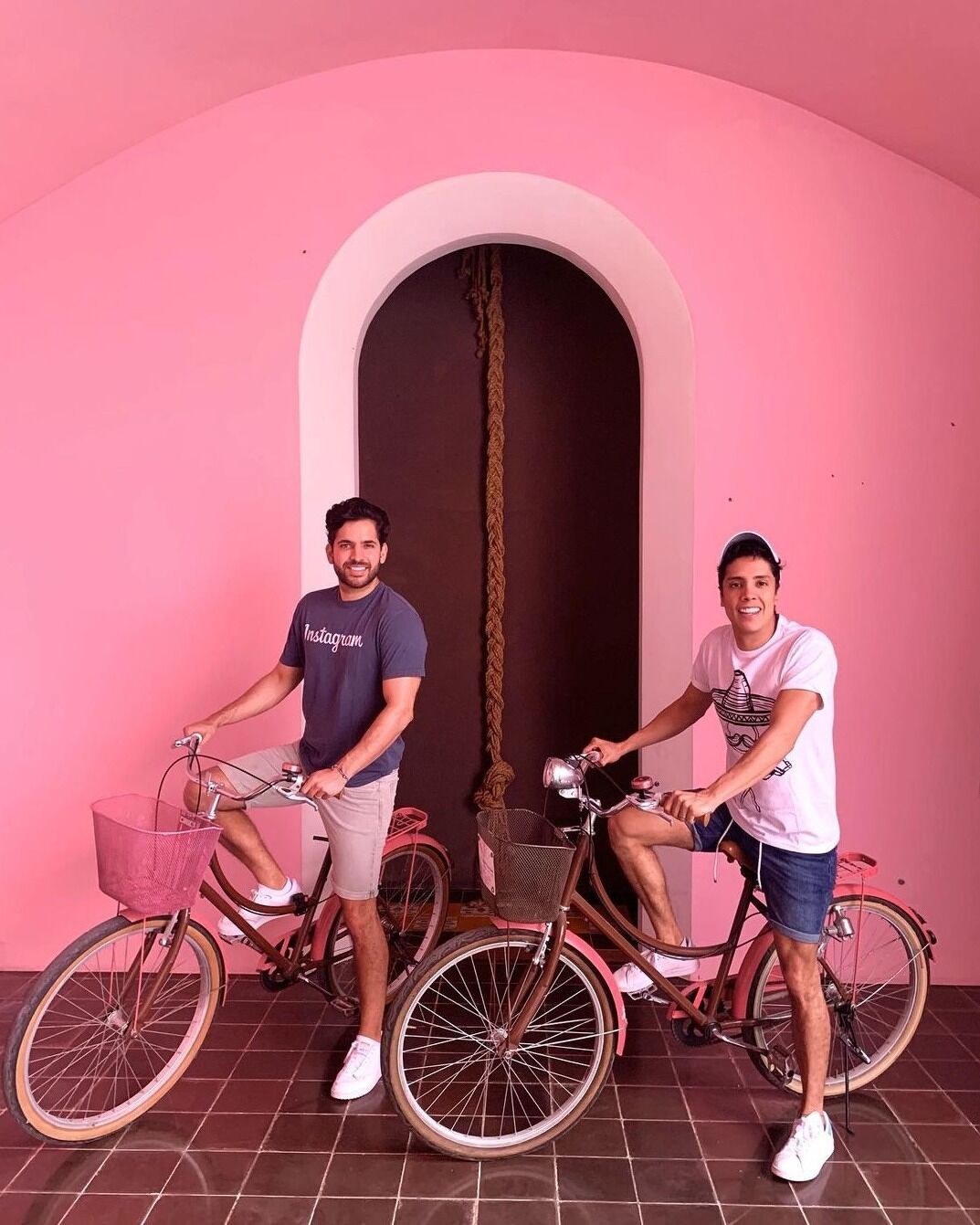 Two handsome men pose with bikes in front of a pink wall.