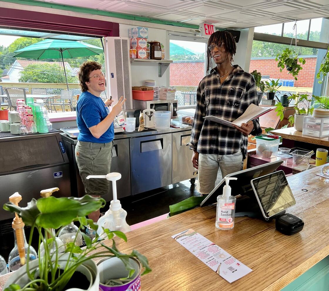 Two baristas, one in a blue shirt and glasses with short curly hair and another in plaid with dreadlocks laugh while making drinks at Finca to Filter cafe in Atlanta. 