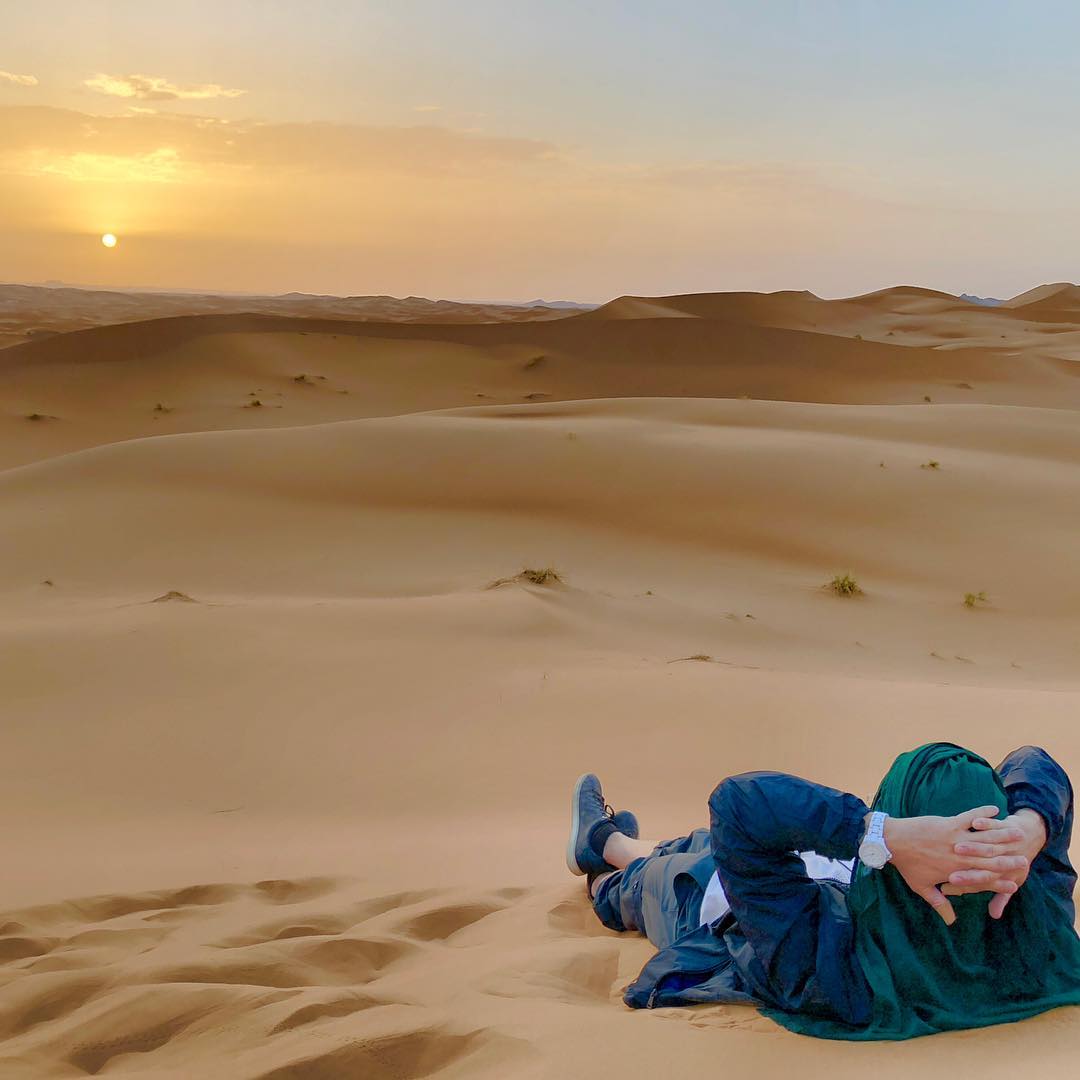 A man rests in the desert to watch the sunset over the sand dunes. 