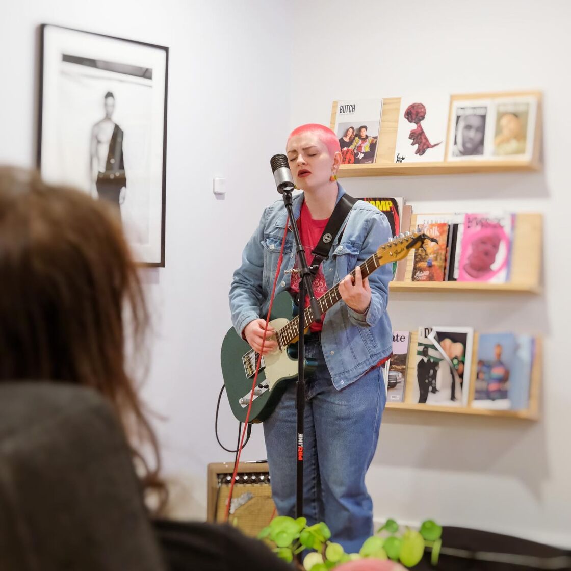 A pink-haired alternative person sings into the mic and plays a green guitar during an open mic night at Modal Coffee and Hostel in Grenville, SC.