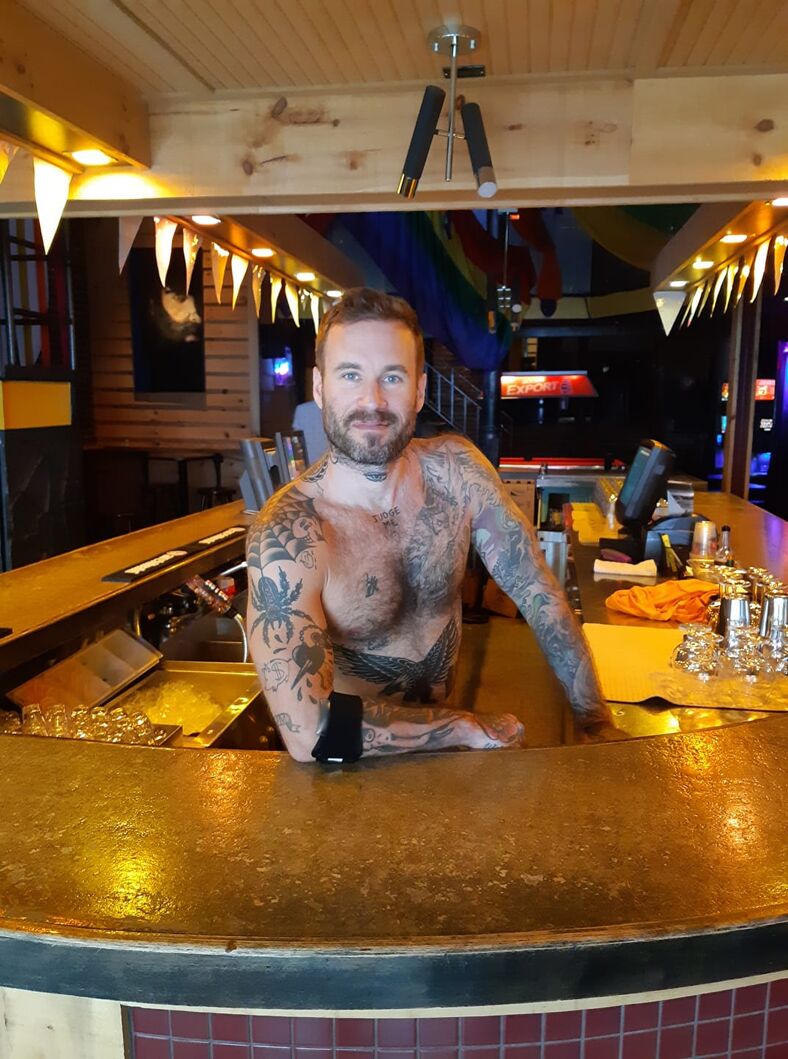 A tattooed, blue-eyed,  muscled, and attractive  shirtless bartender looks ta the camera.