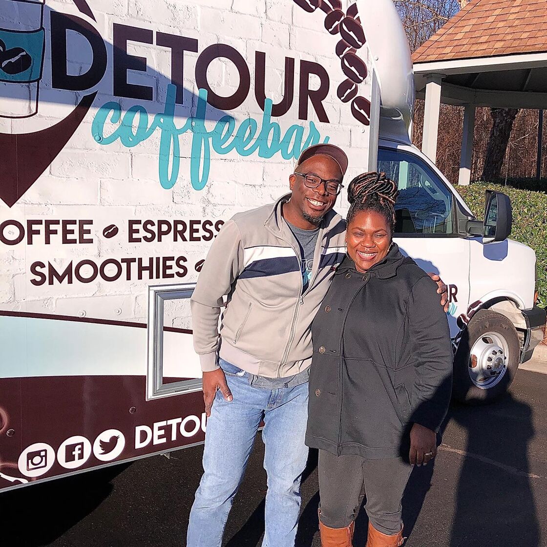 Two smiling black people embrace in front of the Detour Coffeebar truck in Charlotte North Carolina.