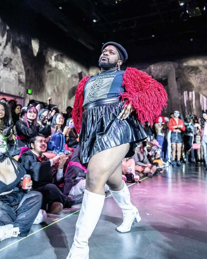 Marco Labeija struts don the catwalk in a punk Joy Division t-shirt, a red furry jacket, black mini skirt,and a beret. Audience members cheer them on.