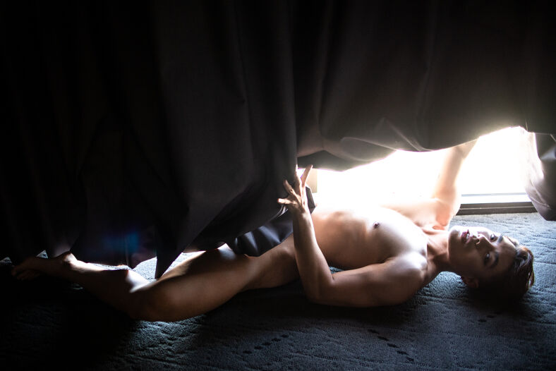 Nude model lying on the floor hides under a curtain, exposing his head, leg and torso for the camera.